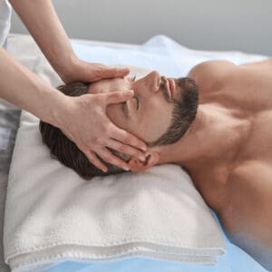 Skilled massage therapist works with head of bearded male client lying on couch with folded towel in contemporary clinic office side view